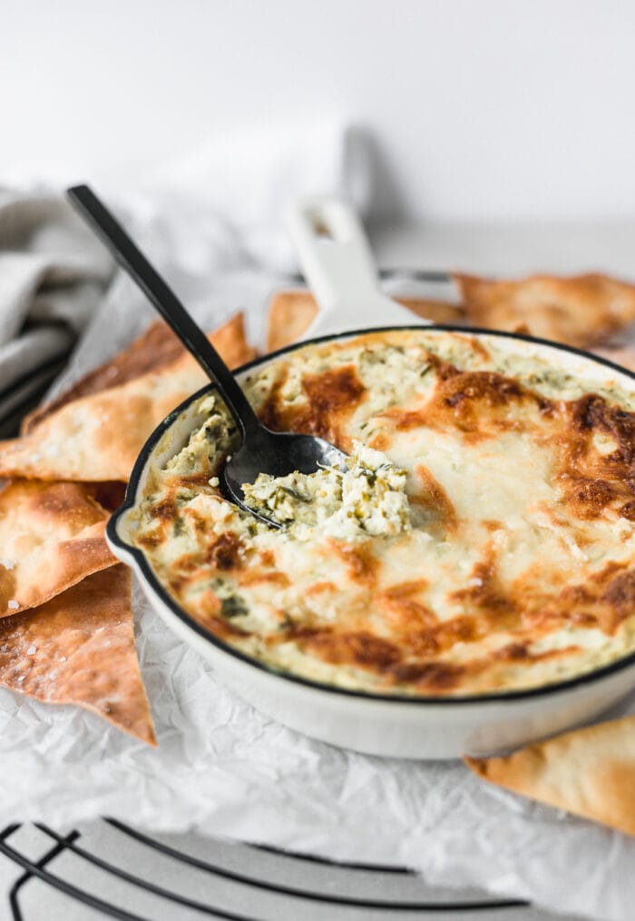 kale artichoke dip in a white skillet with a black spoon inside it.