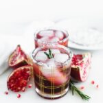 two pomegranate ginger smash cocktails in a straight line in gold plaid cocktail glasses surrounded by pomegranate pieces and rosemary.