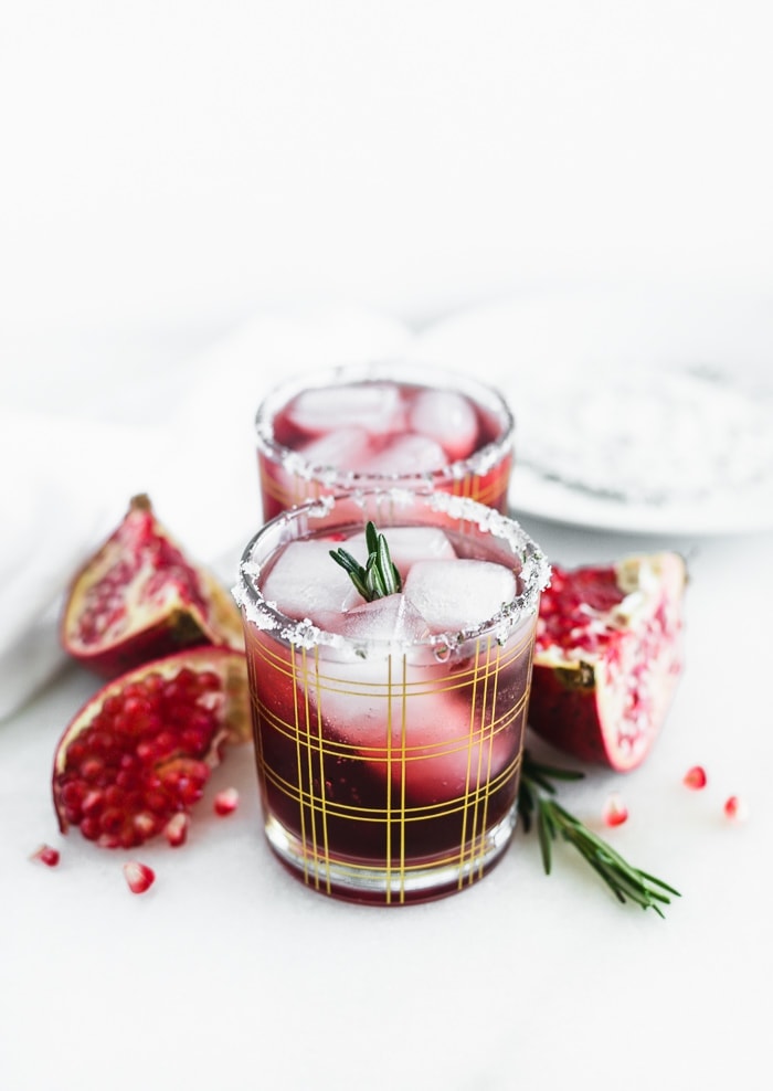 two pomegranate ginger smash cocktails in a straight line in gold plaid cocktail glasses surrounded by pomegranate pieces and rosemary.
