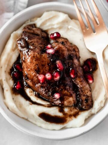pomegranate balsamic chicken thigh on top of mashed potatoes in a grey bowl with a gold fork.