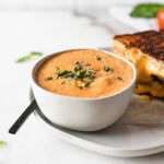 Roasted Red Pepper Tomato Basil Soup sprinkled with basil in a small bowl next to a grilled cheese sandwich.