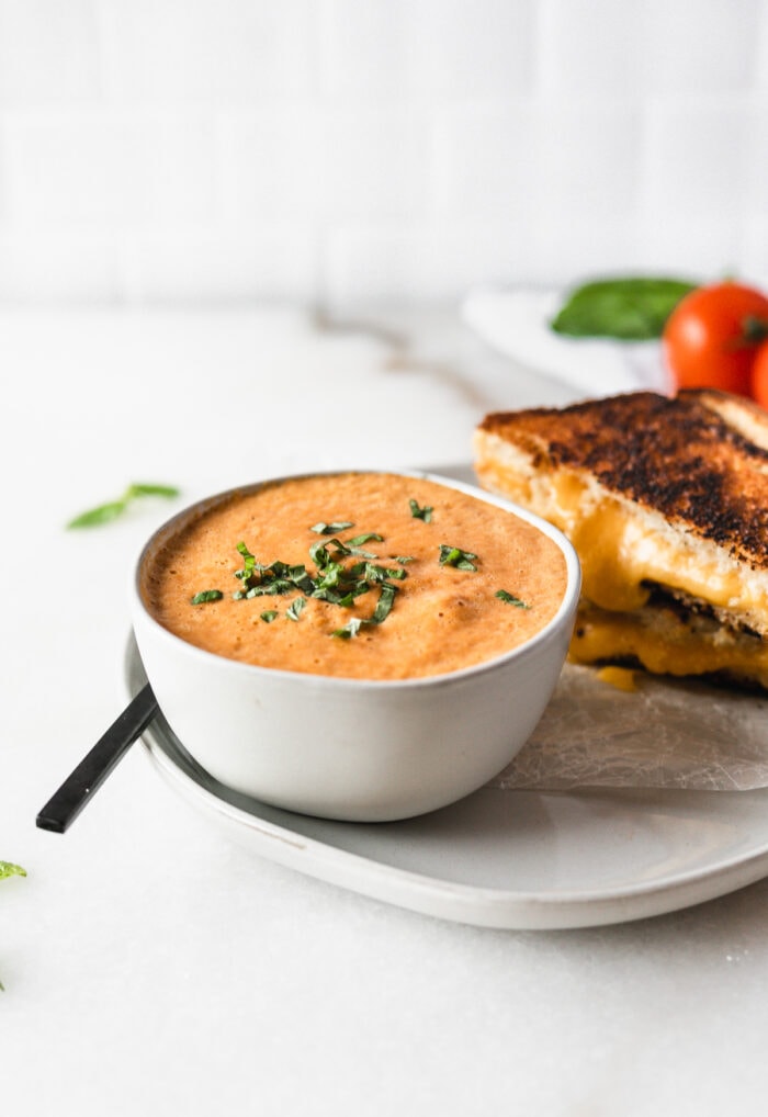 Roasted Red Pepper Tomato Basil Soup sprinkled with basil in a small bowl next to a grilled cheese sandwich.
