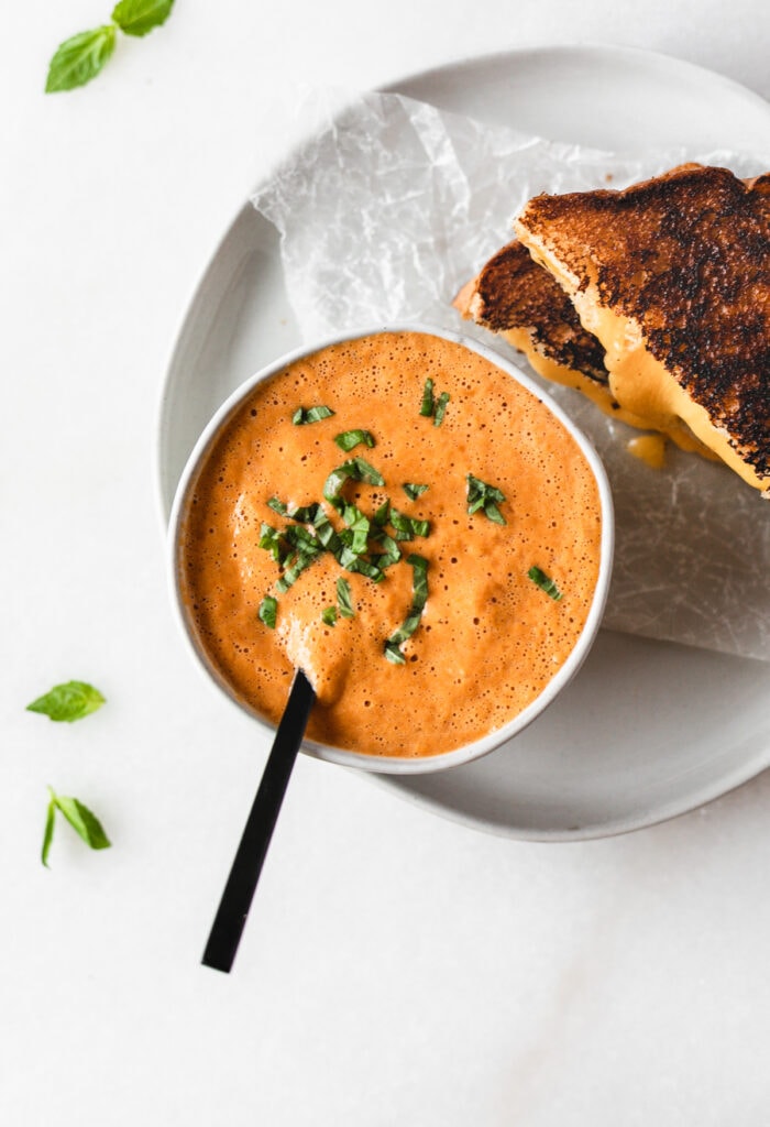 Overhead view of Roasted Red Pepper Tomato Basil Soup sprinkled with basil in a small bowl with a black spoon in it next to a grilled cheese sandwich on a plate.