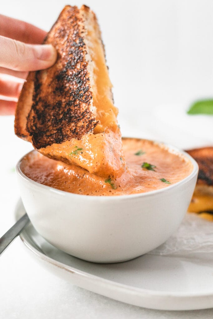 Half a grilled cheese sandwich being dipped into a bowl of Roasted Red Pepper Tomato Basil Soup.