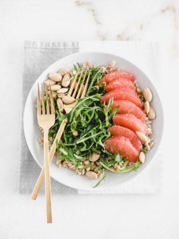 overhead view of grapefruit arugula farro salad in a white bowl with gold forks on top of a gray and white napkin.
