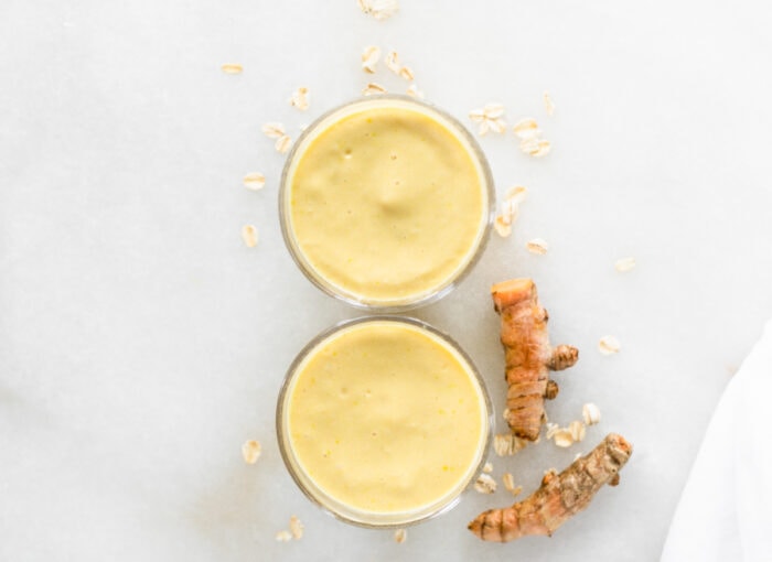 overhead view of 2 mango ginger turmeric smoothies on a white background with oats and turmeric roots around them.