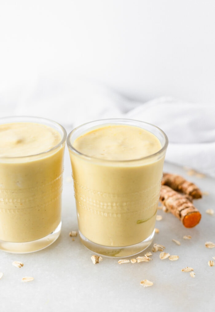 closeup of mango ginger turmeric smoothie in a glass with oats and turmeric root scattered around it.