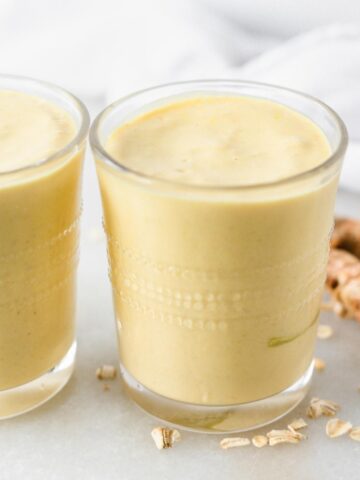 closeup of mango ginger turmeric smoothie in a glass with oats and turmeric root scattered around it.
