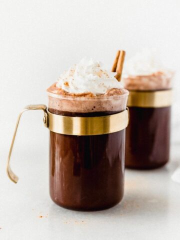 closeup of a glass mug of tequila spiked Mexican hot chocolate topped with whipped cream and a cinnamon stick.
