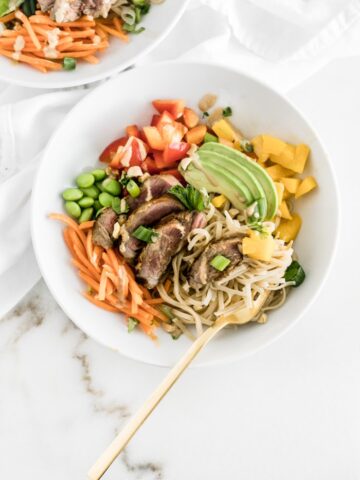 overhead view of Thai mango steak noodle bowl in white bowl with a gold fork in it and a white napkin beside it.