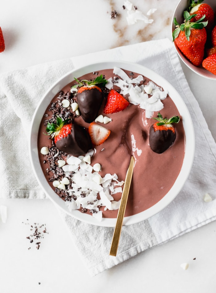 overhead view of chocolate strawberry smoothie bowl in a white bowl with chocolate covered strawberries and coconut shavings on top with a gold spoon in it.