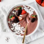 overhead view of chocolate strawberry smoothie bowl in a white bowl with chocolate covered strawberries and coconut shavings on top with a gold spoon in it.