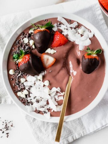 overhead view of chocolate strawberry smoothie bowl in a white bowl with chocolate covered strawberries and coconut shavings on top with a gold spoon in it.