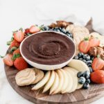 brownie batter dip in a white bowl on a round wooden board with fruit, cookies, and pretzels.