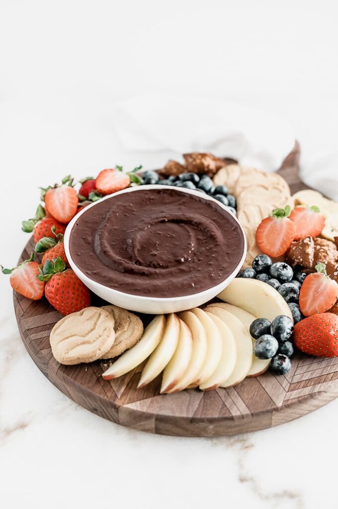 brownie batter dip in a white bowl on a round wooden board with fruit, cookies, and pretzels.
