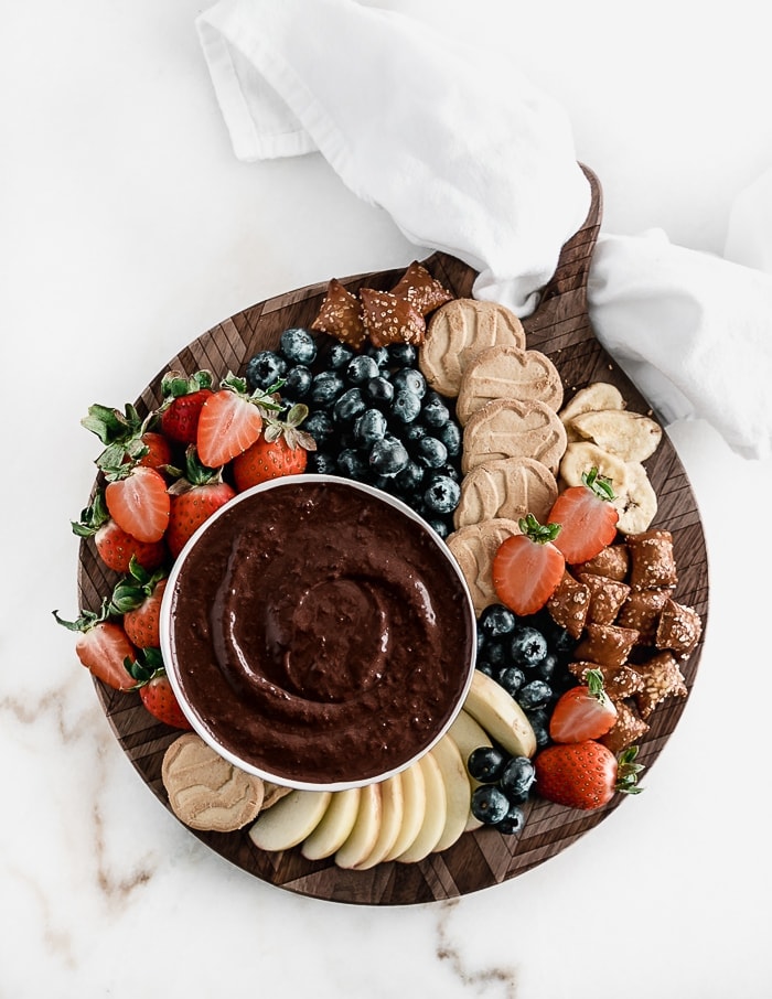 overhead shot of brownie batter dip in a white bowl on a round wooden board with fruit, cookies, and pretzels.