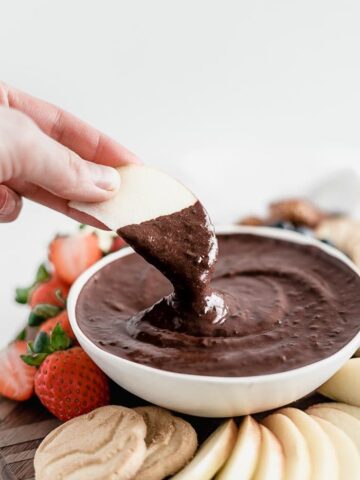 hand dipping an apple slice into a bowl of brownie batter dip surrounded by cookies, strawberries and apples.