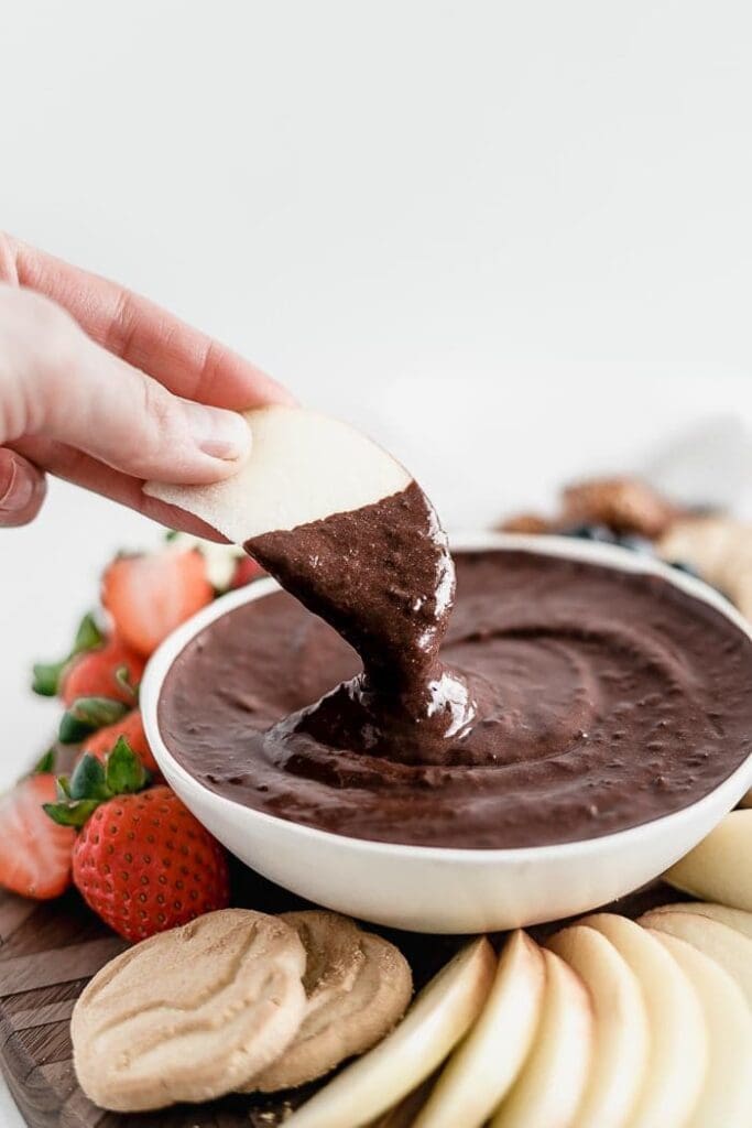 hand dipping an apple slice into a bowl of brownie batter dip surrounded by cookies, strawberries and apples.