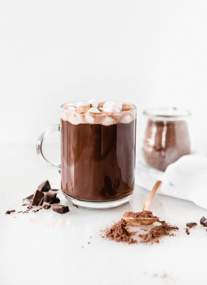 mug of homemade dark hot cocoa topped with marshmallows with a gold spoon of cocoa mix, dark chocolate chunks, and a jar of the mix surrounding it.