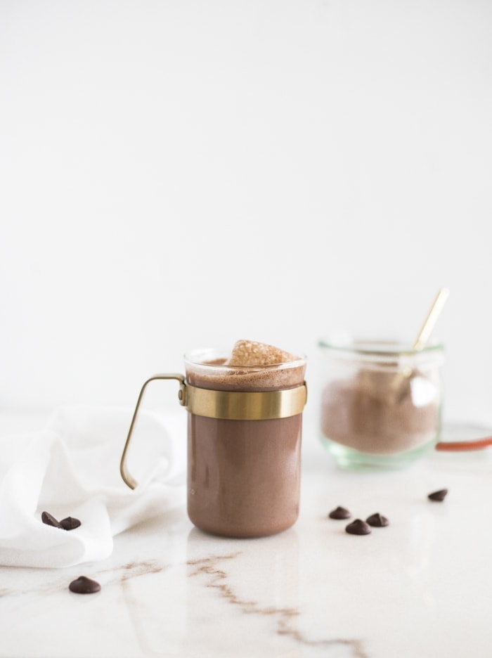 mug of homemade dark hot cocoa topped with marshmallows with a gold spoon of cocoa mix, dark chocolate chunks, and a jar of the mix surrounding it.