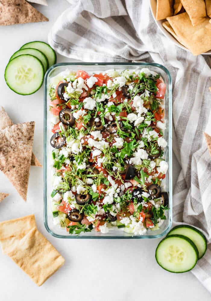 overhead view of 7 layer greek dip in a glass dish with a grey and white striped napkin, pita wedges, pita chips and cucumber slices around it.