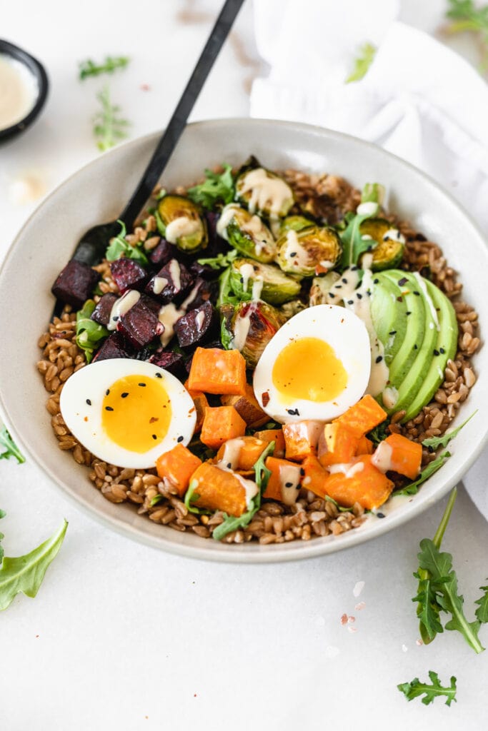 roasted veggie grain bowl with farro, roasted sweet potato, beets, brussels sprouts, and avocado topped with a soft boiled egg and tahini sauce with a black fork in it.