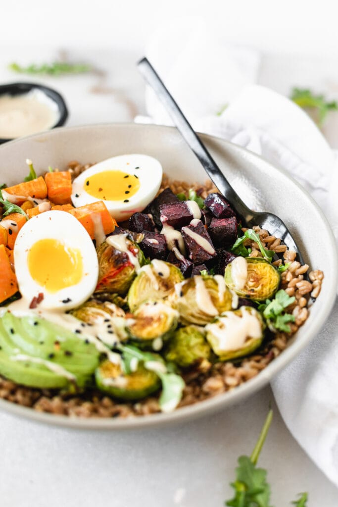 closeup of a grain bowl with farro, sweet potato, beets, brussels sprouts, a soft boiled egg and tahini sauce.