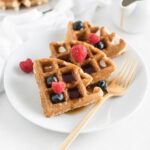 three sourdough waffle triangles on a white plate topped with berries and syrup with a gold fork beside them.