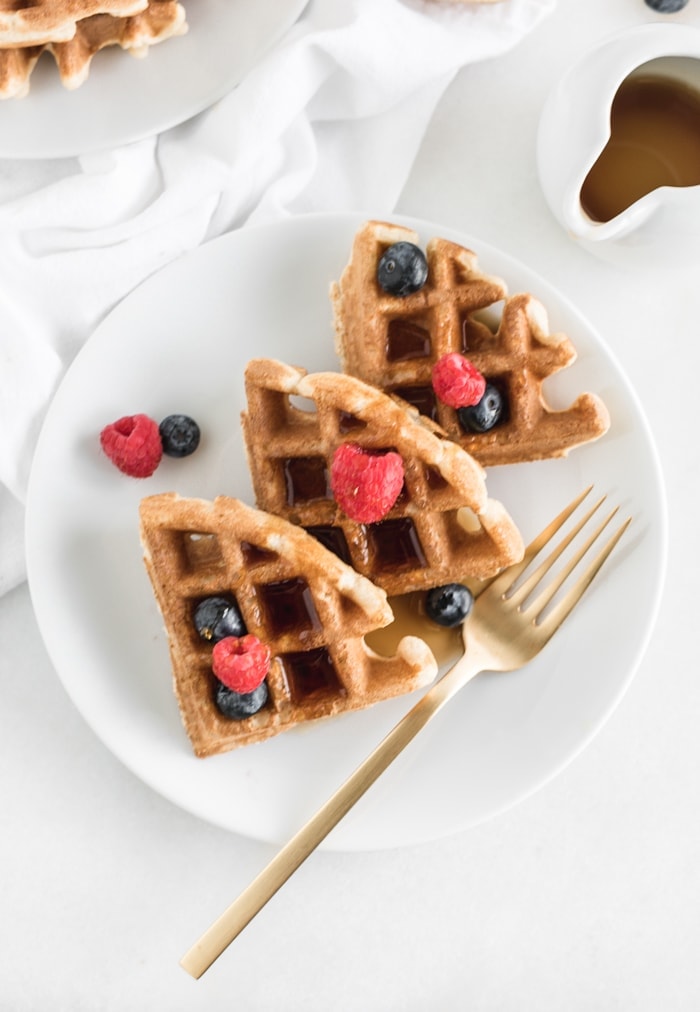 overhead view of three sourdough waffle triangles on a white plate topped with berries and syrup with a gold fork beside them.