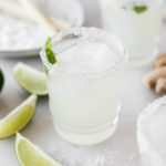 lemongrass margarita in a glass with cilantro and ice in it surrounded by lime slices and salt.