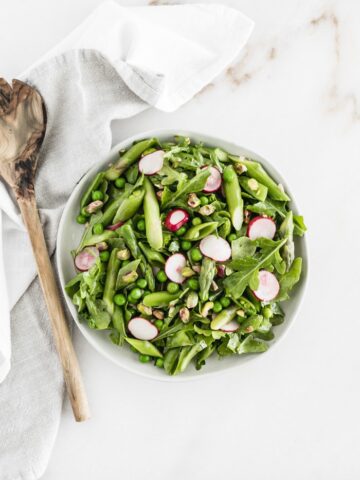 Overhead view of arugula spring salad on a white plate with a wooden serving spoon and gray napkin next to it.
