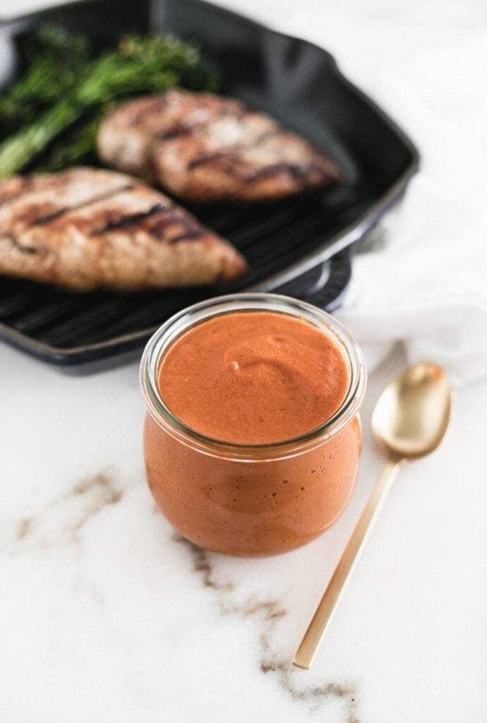 homemade romesco sauce in a glass jar with a gold spoon next to it and a grill pan with chicken in the background.