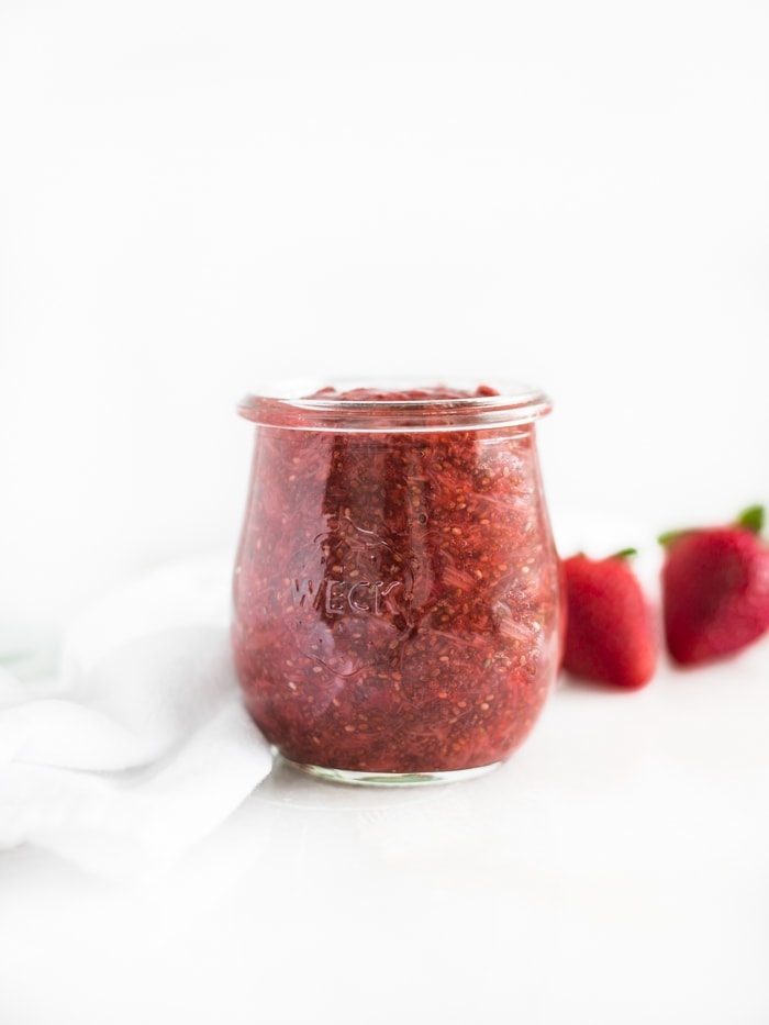 Strawberry rhubarb chia jam in a clear jar with a white napkin and fresh strawberries next to it.