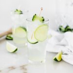 white linen cocktail in a glass garnished with lime and cucumber surrounded by lime wedges.