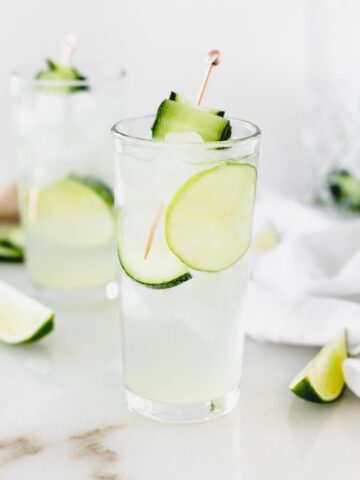 white linen cocktail in a glass garnished with lime and cucumber surrounded by lime wedges.