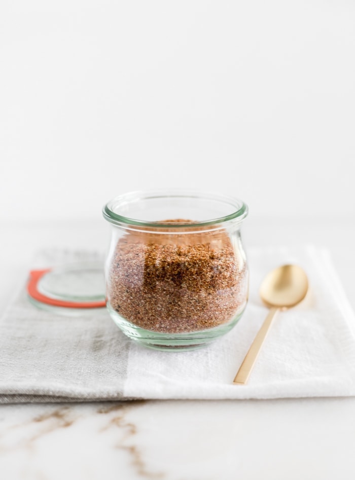 BBQ spice rub in a glass jar with a gold spoon beside it on top of a grey and white napkin.