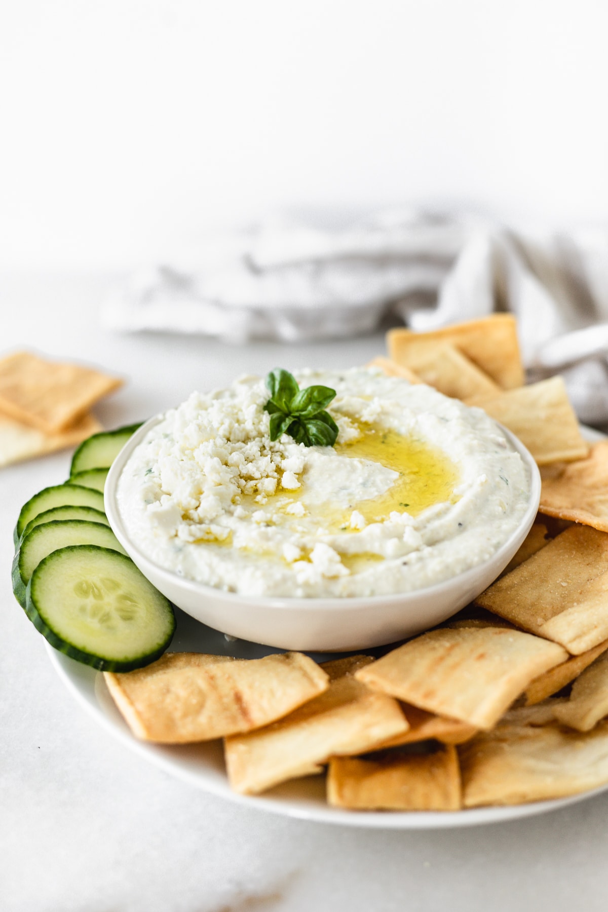 whipped feta artichoke dip in a white bowl topped with feta, basil and olive oil surrounded by pita chips and sliced cucumbers.