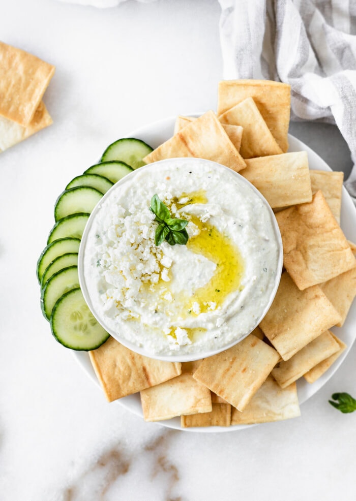 overhead view of whipped feta artichoke dip in a bowl surrounded by pita chips and sliced cucumbers.