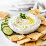 whipped feta artichoke dip in a white bowl topped with feta, basil and olive oil surrounded by pita chips and sliced cucumbers.