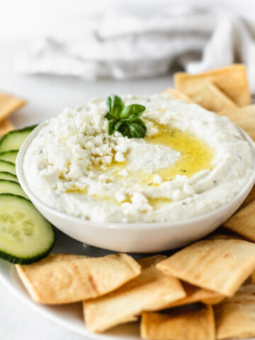 whipped feta artichoke dip in a white bowl topped with feta, basil and olive oil surrounded by pita chips and sliced cucumbers.