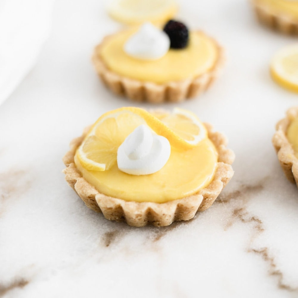 mini lemon tart with shortbread crust with whipped cream and a lemon slice on top, with lemon tarts in the background.