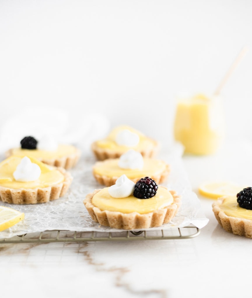 mini lemon tarts topped with whipped cream, berries, and lemon slices on a cooling rack.