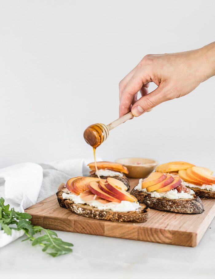 hand drizzling honey over peach bruschetta with a honey dipper.