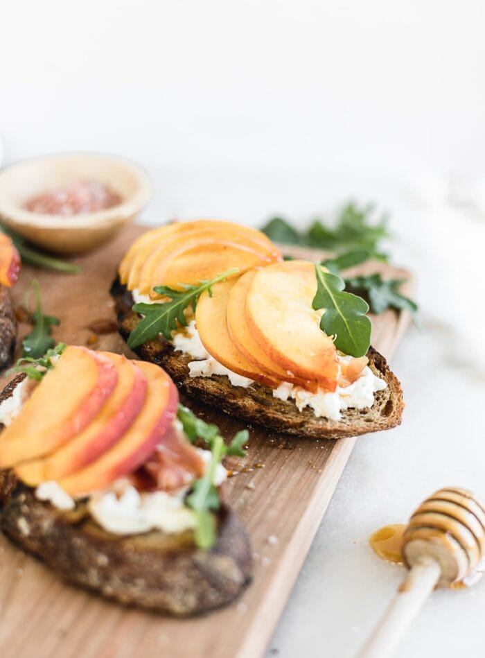 Piece of peach bruschetta on a cutting board surrounded by arugula a bowl of salt and a honey dipper.