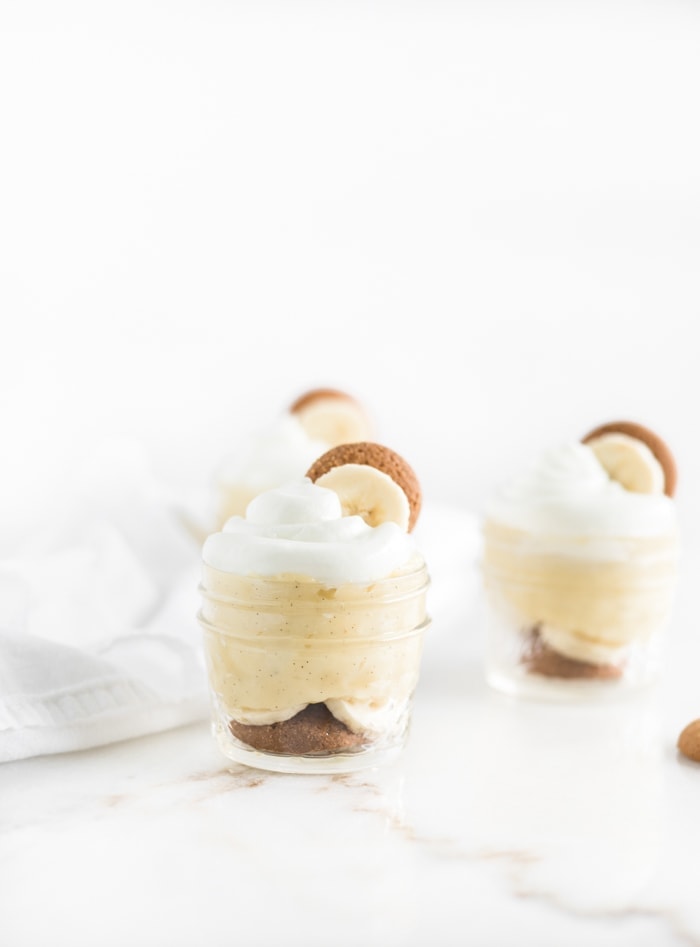 3 small glass jars of banana pudding with whipped cream, a banana slice and a vanilla wafer on top.