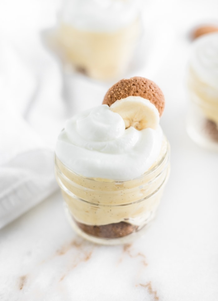small glass jar of banana pudding with whipped cream, a banana slice and a vanilla wafer on top.