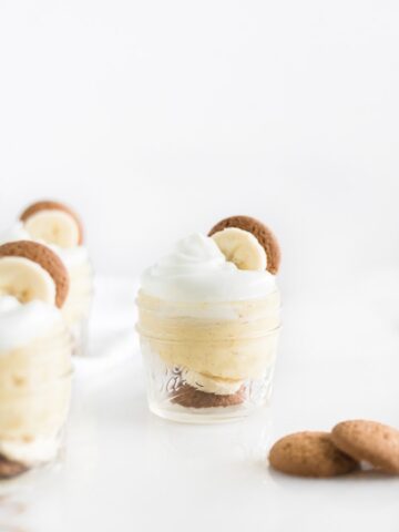 small glass jar of banana pudding with whipped cream, a banana slice and a vanilla wafer on top, surrounded by more jars of pudding and vanilla wafers.