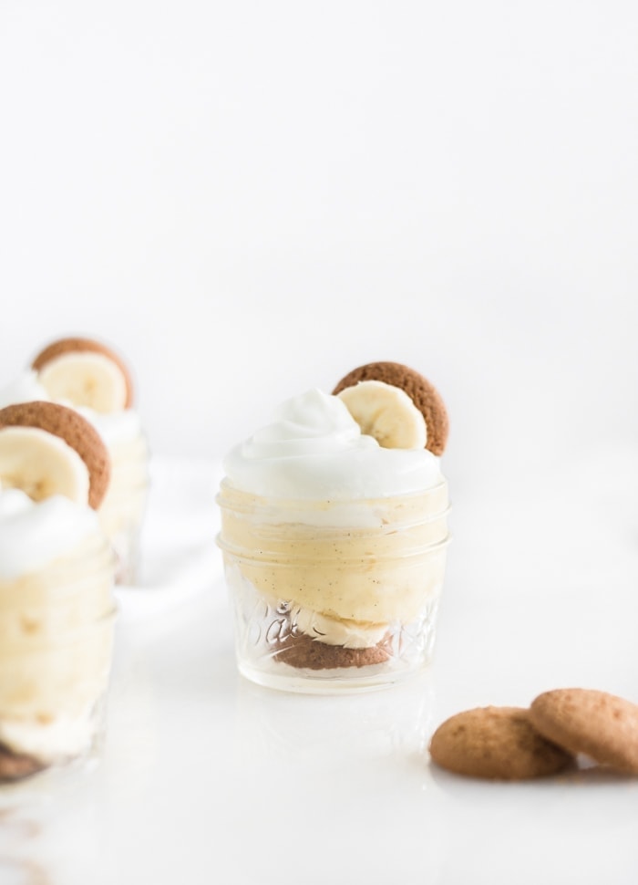 small glass jar of banana pudding with whipped cream, a banana slice and a vanilla wafer on top, surrounded by more jars of pudding and vanilla wafers.
