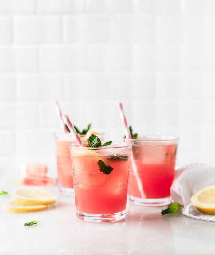 three glasses of watermelon mint lemonade with pink striped straws surrounded by lemon slices and mint leaves.