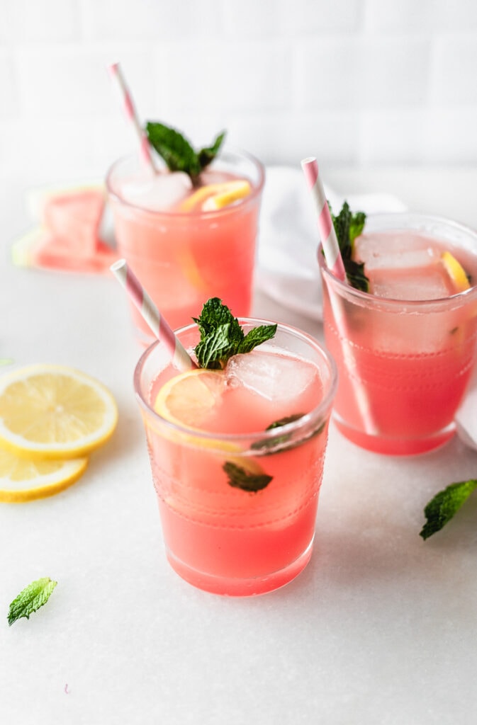 glass of sparkling watermelon mint lemonade with a pink striped straw with two more glasses behind it.